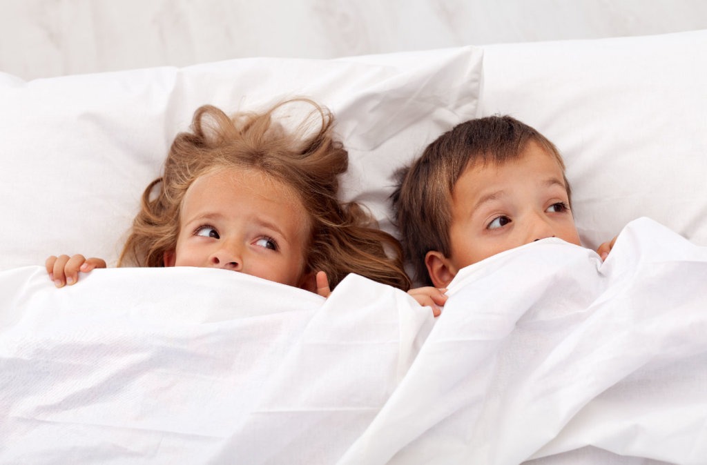 Duas crianças deitadas na cama, parcialmente cobertas, com expressões de medo, ilustrando os medos noturnos comuns, como o medo do escuro e de monstros.