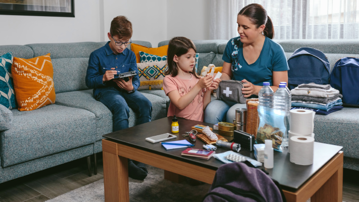 Família com crianças a preparar kit de emergência em casa, com água, alimentos e artigos de higiene sobre a mesa